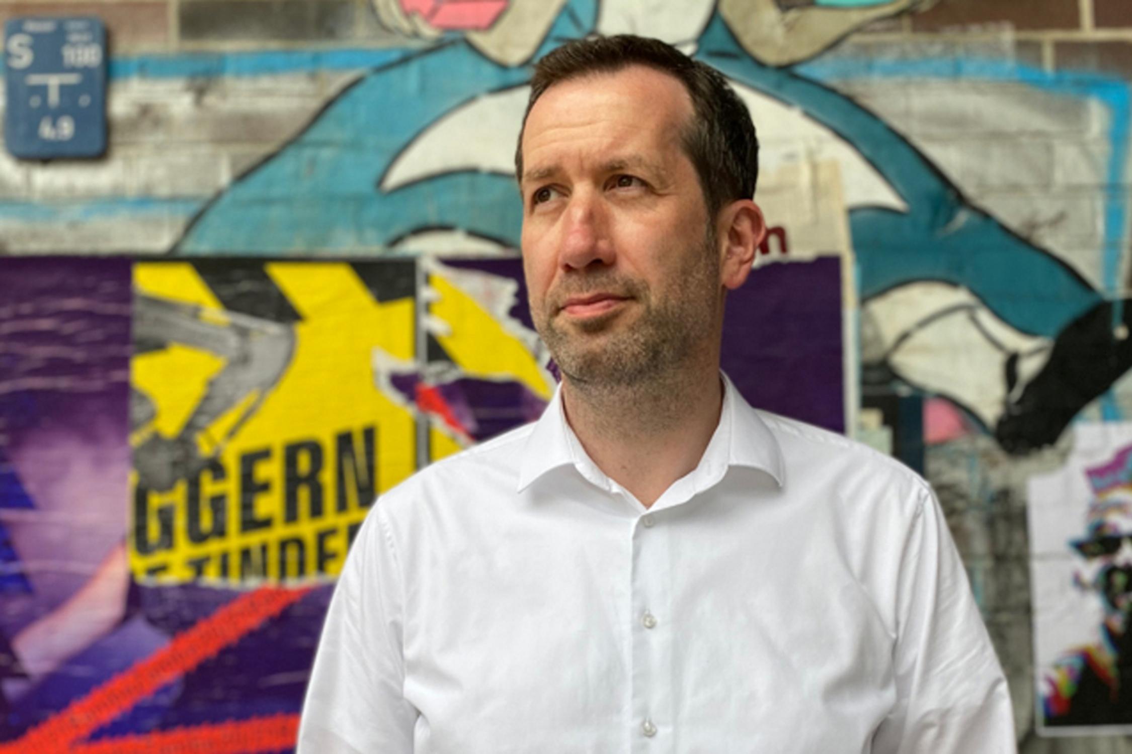 Ein Mann im Portrait steht vor einer Graffiti-Mauer und schaut schräg nach links oben.