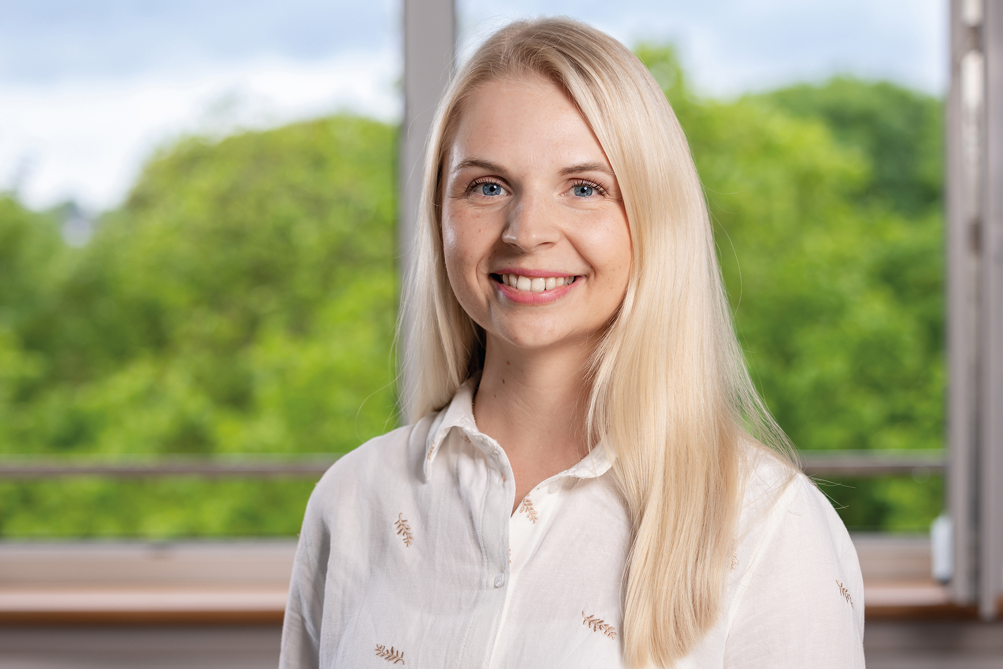 Eine Frau mit langen blonden Haaren lächelt in die Kamera und steht vor einem Fenster.