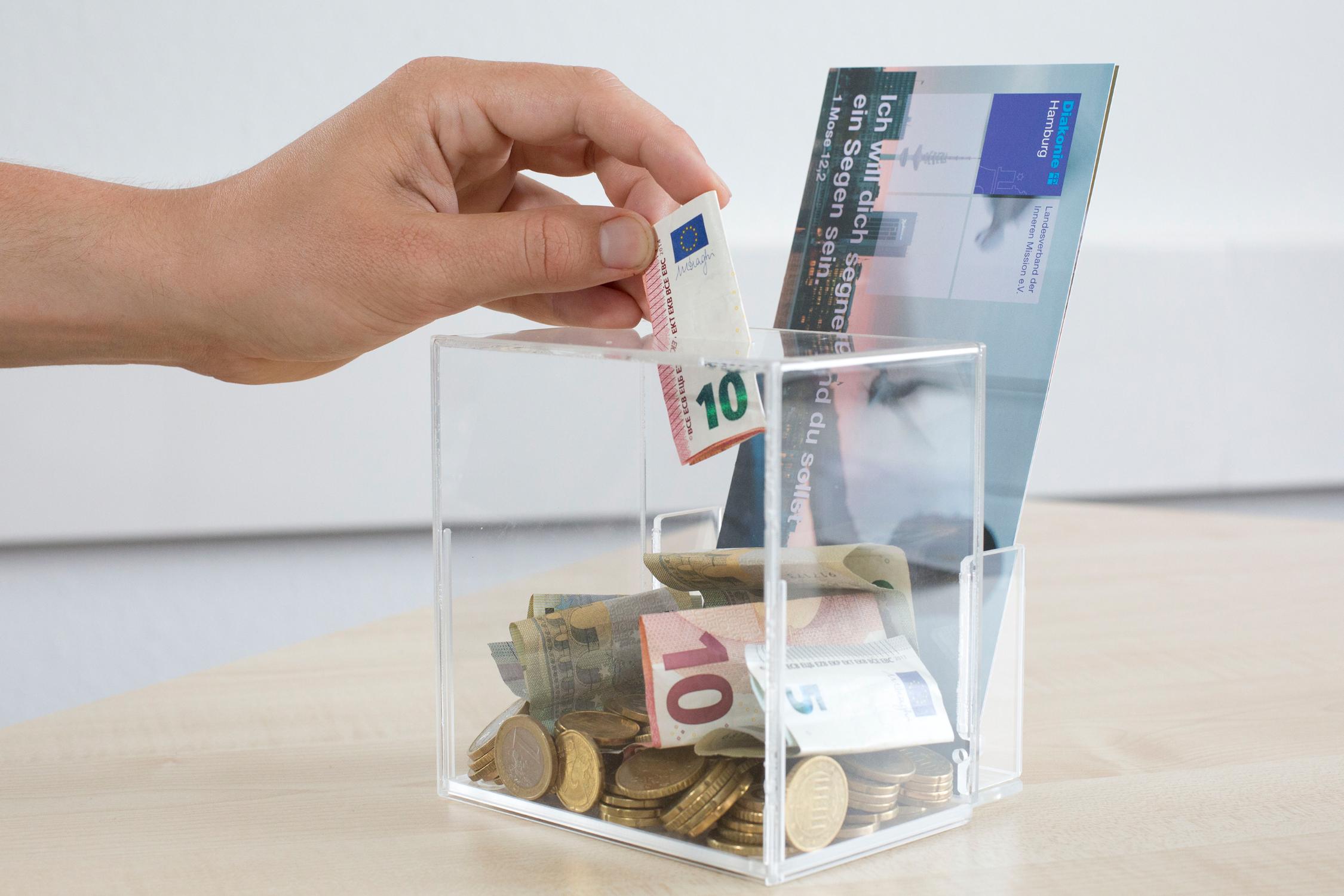Eine Hand wirft einen Zehn-Euro-Schein in eine durchsichtige Spardose, in der bereits Geldscheine und Münzen liegen.